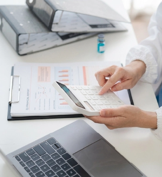 An accountant using a white calculator over financial reports and a laptop on a white desk.