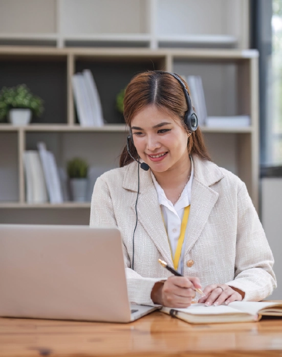 A smiling support representative with a headset taking notes while working on a laptop.