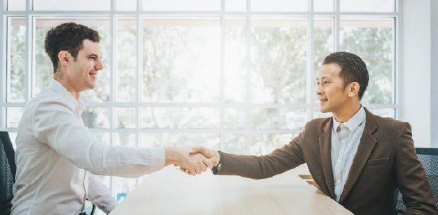 Two businessmen in a bright office sitting at a desk and shaking hands with friendly expressions.